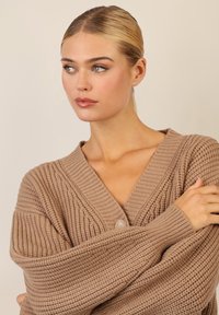 Cardigan chunky a maglia in marrone chiaro, con scollo a V e un grande bottone. Tessuto strutturato con maniche ampie e dettagli a coste.