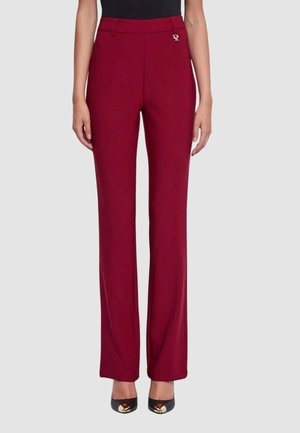 Donna che indossa pantaloni bordeaux a vita alta e gamba dritta con passanti per cintura e piccolo logo dorato, abbinati a tacchi neri e top nero.