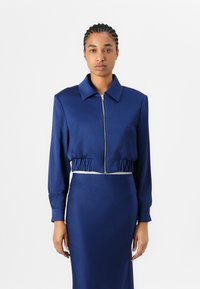 Giacca cropped blu navy con zip frontale, colletto e vita elasticizzata, abbinata a una gonna aderente coordinata. Texture del tessuto liscia.