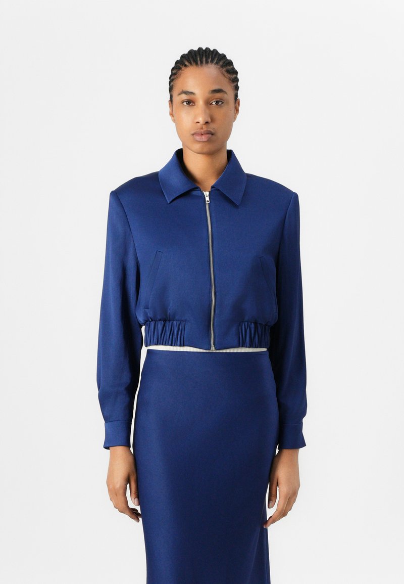 Giacca cropped blu navy con zip frontale, colletto e vita elasticizzata, abbinata a una gonna aderente coordinata. Texture del tessuto liscia.