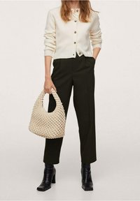 Crèmekleurig vest met ronde hals en knoopsluiting, gecombineerd met donkergroene broek en zwarte laarzen met hak, met een geweven beige tas in de hand.