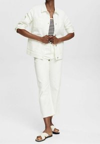 Person som bär en vit denimjacka och byxor, topp med svart-vita ränder och vita slip-on sandaler, står framför en enfärgad bakgrund.