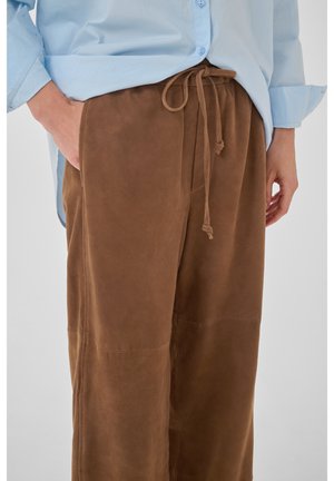 Hele sinine pikkade varrukatega särk, pistetud pruunidesse seemisnahast nöörpükstesse, üks käsi taskus ja teine lõdvestunult kõrval.