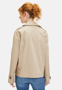Beige trench coat with a classic cut, featuring shoulder epaulettes, buttoned cuffs, and a smooth texture, paired with blue jeans.