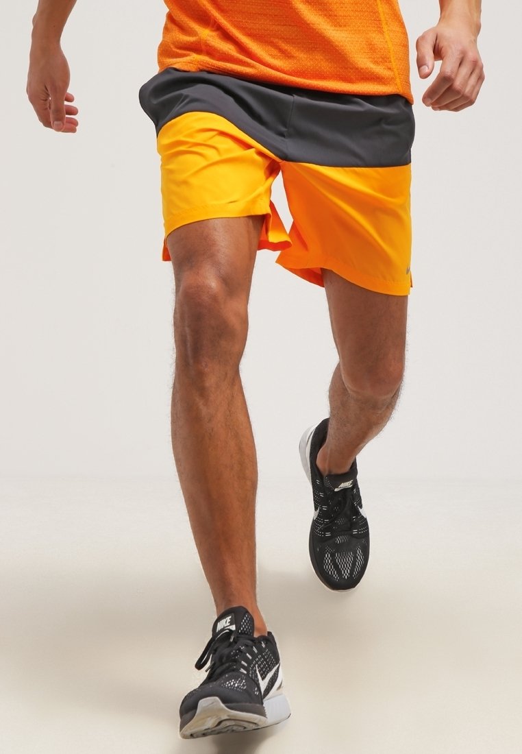 Homme courant portant un short orange et noir, un t-shirt orange et des chaussures de course noires Nike sur un fond clair.
