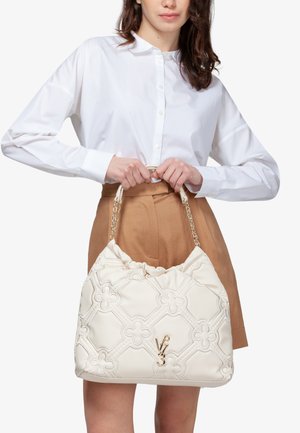 Witte gewatteerde handtas met bloemenpatroon, gouden kettingaccenten en geplooide bovenkant. Model houdt de tas vast, gekleed in een witte blouse en een beige rok.