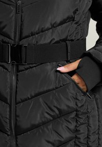 Schwarze Daunenjacke mit gestepptem Design, flexibler Gurt mit glänzender Schnalle und Reißverschlusstaschen. Weicher, strukturierter Stoff.