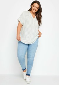 Blusa leggera a righe color crema con scollo a V e maniche corte, abbinata a jeans skinny blu chiaro e sneakers bianche.