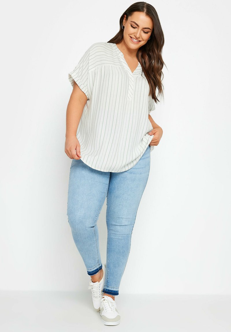 Blusa leggera a righe color crema con scollo a V e maniche corte, abbinata a jeans skinny blu chiaro e sneakers bianche.