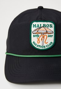 Gorra negra con visera curvada, que presenta un parche bordado en verde y blanco de un paraguas y el texto "MALBON VACATION CLUB". Ribete de acento verde.