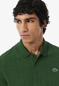 Polo verde realizzato in tessuto testurizzato con un sottile motivo a griglia. Presenta un colletto, tre bottoni e un piccolo logo sul petto.