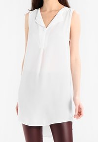 Blusa blanca sin mangas con cuello tipo solapa y una fila de botones, hecha de una tela ligera. La parte trasera es ligeramente más larga que la delantera.