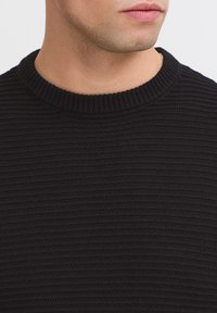 Zwart gebreid sweater met een ronde hals, met een getextureerd ribpatroon en elasticiteit. Close-up van de halslijn en het bovenste borstgebied.