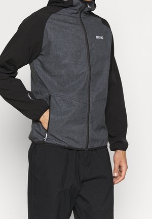 Mann trägt eine schwarz-graue Kapuzenjacke mit Reißverschluss, die Hand in der Tasche, kombiniert mit schwarzer Hose vor einfarbig hellem Hintergrund.