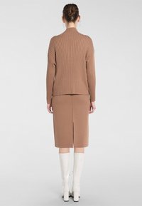 Pull en maille côtelée marron avec un col montant, associé à une jupe marron longueur genoux avec une fente arrière, portée avec des bottes blanches à genoux.