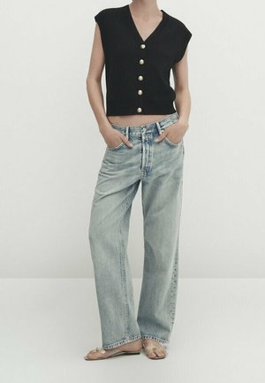 Black sleeveless cropped top with gold buttons, paired with light blue high-waisted straight-leg jeans. Barefoot and hands in pockets.