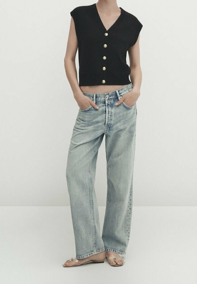 Black sleeveless cropped top with gold buttons, paired with light blue high-waisted straight-leg jeans. Barefoot and hands in pockets.