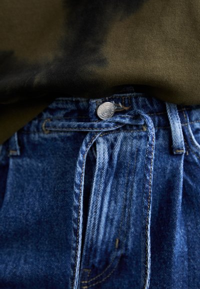 Blugi din denim cu o spălare albastră, dotati cu închidere cu button metalic și șnur la talie, asortați cu un tricou de culoare verde închis cu efect tie-dye.