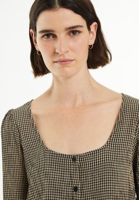 Blusa de manga larga con escote cuadrado, patrón de cuadros en negro y beige, y cierre de botones negros en la parte delantera. Tejido texturizado.