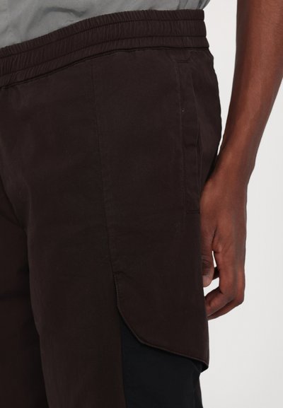 Pantalons marron foncé avec une taille élastique, dotés d'une poche latérale et d'un tissu légèrement texturé. Coutures visibles le long de la surface.