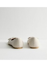 Ballerine in tessuto beige con finitura texturizzata, punta arrotondata e un fiocco sul retro. Suola piatta con un bordo marrone chiaro.