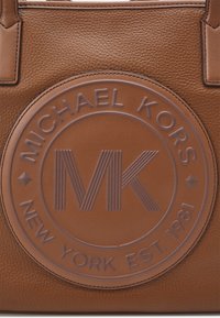 Borsa a mano in pelle marrone con un logo circolare in rilievo riportante il testo "MICHAEL KORS" e "NEW YORK EST. 1981" e un design prominente "MK".