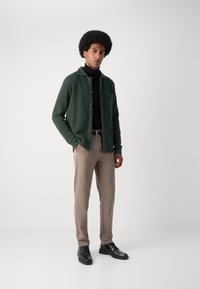 Cárdigan de punto verde con botones marrones, bolsillo, llevado sobre un cuello de tortuga negro. Pantalones beige y zapatos negros de cordones completan el conjunto.