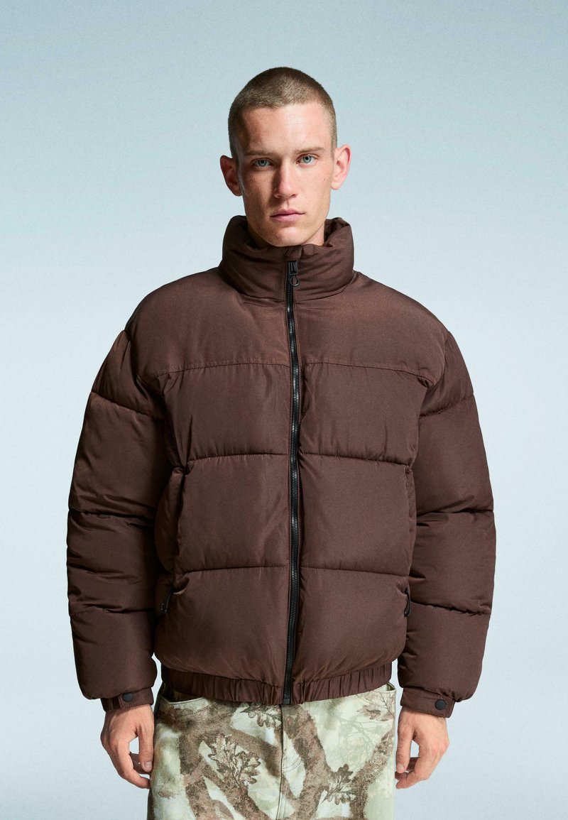 Giubbotto imbottito marrone con colletto alto, presenta un motivo trapuntato, chiusura a zip e orlo elasticizzato. Indossato con pantaloni chiari a fantasia.