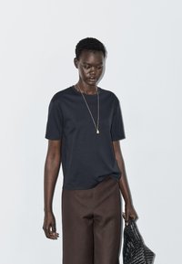 Mørkeblå t-shirt med korte ærmer lavet af glat stof, kombineret med brune løstsiddende bukser. Inkluderer guldfarvet vedhængshalskæde og grå vævet taske.