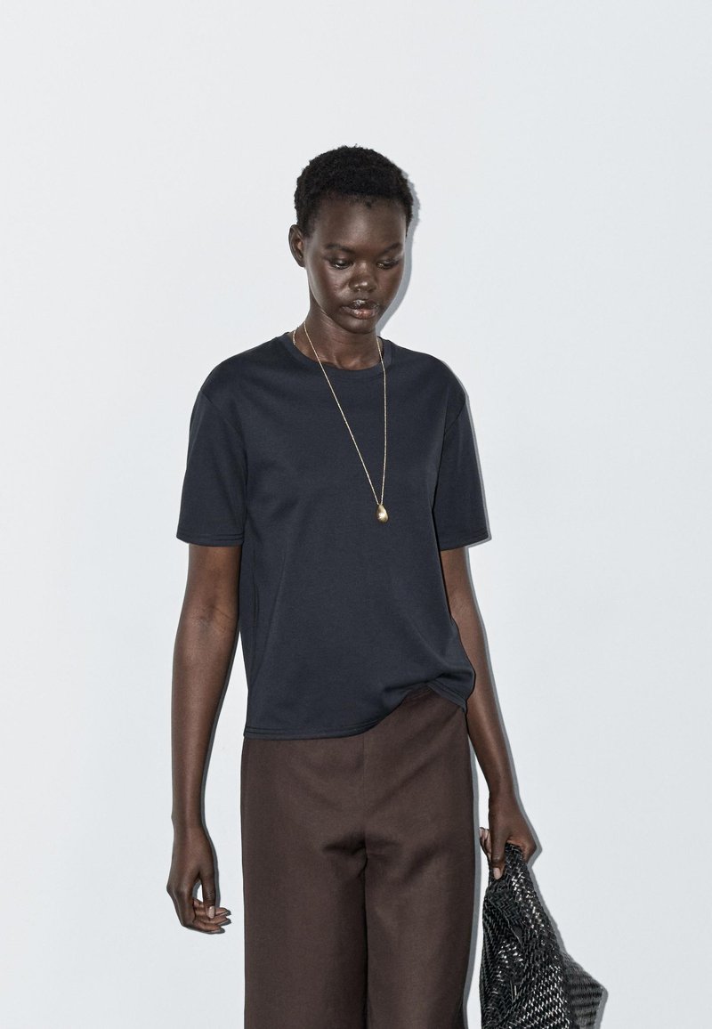 Mørkeblå t-shirt med korte ærmer lavet af glat stof, kombineret med brune løstsiddende bukser. Inkluderer guldfarvet vedhængshalskæde og grå vævet taske.