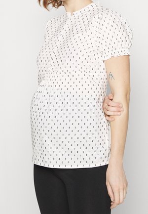 Blusa ligera de color blanco con un patrón de puntos negros, mangas cortas y abullonadas, y cintura fruncida. Tejido suave con un cuello.