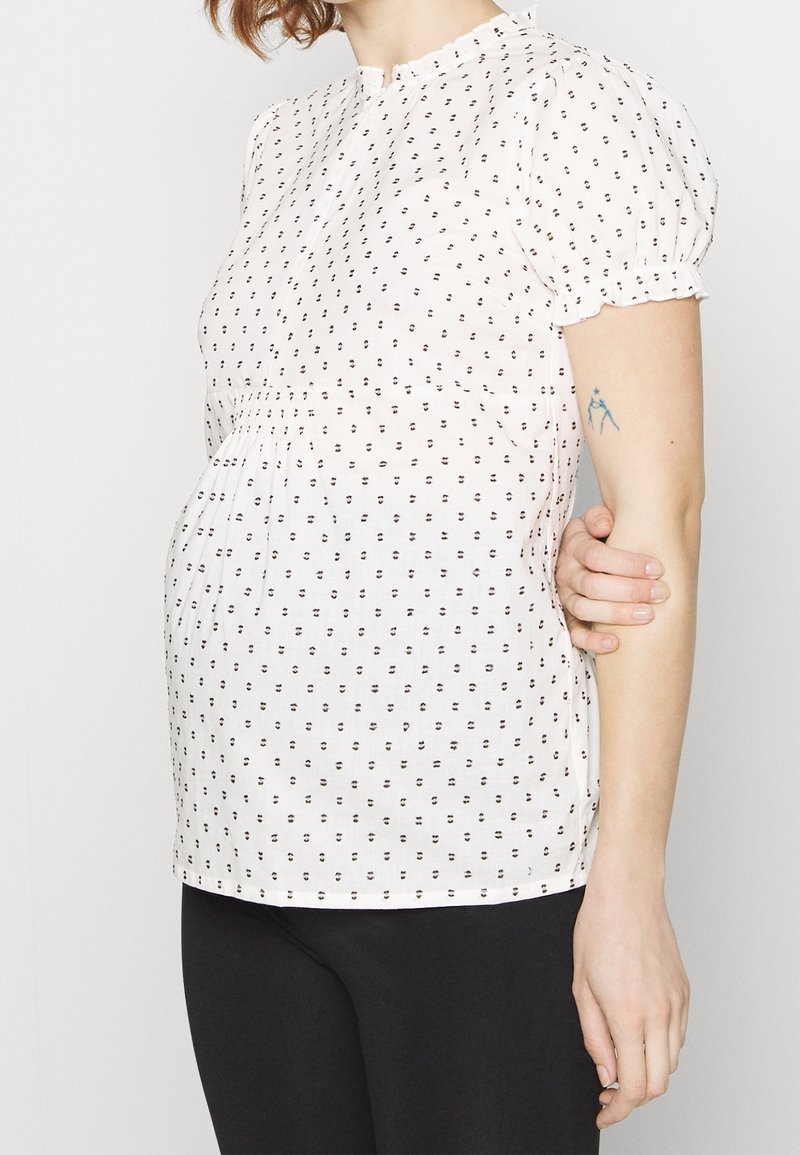 Blouse légère, blanche avec un motif de points noirs, à manches courtes bouffantes et taille froncée. Tissu lisse avec un col.