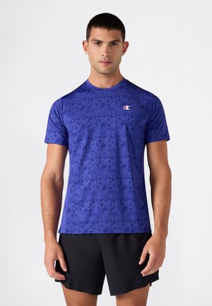 Man met een blauw sportshirt met korte mouwen en patroon en zwarte korte broek, staand tegen een effen witte achtergrond.