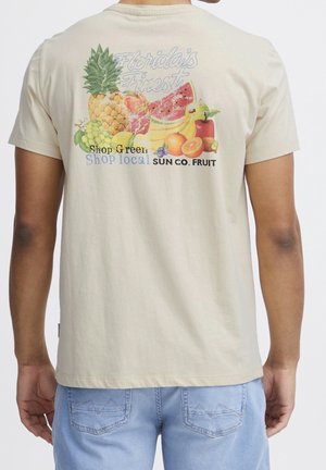 Mann trägt beiges T-Shirt mit bunter Obstgrafik und Text über Florida und lokales Einkaufen auf der Rückseite, kombiniert mit hellblauen Jeans.