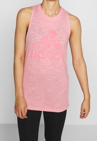 Světle růžový sportovní top bez rukáv�ů s kulatým výstřihem a vybledlým černým logem Adidas na přední straně. Měkká, strukturovaná látka.