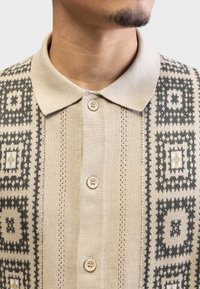 Beige strikket polo med et geometrisk mønster i mørkegrått og kremfarge, med klassisk krage og to-knapps knapping.