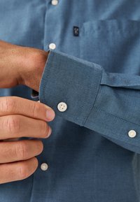 Chemise bleu à boutons en tissu texturé, avec un bouton blanc sur le poignet et une poche poitrine avec un logo discret.