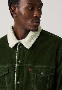 Dark green corduroy jacket with a cream faux shearling collar, front pockets, and metal button closures. Features a red brand tag.