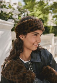 Brun leopardmönstrad pannband i konstpäls och matchande muddar, båda bärs med en svart läderjacka, kännetecknas av en mjuk textur och djärvt mönster.