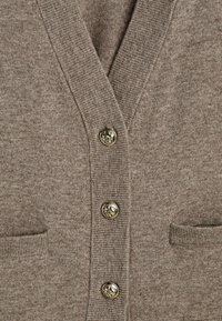 Beige neulejakku, jossa on ribattu V-pääntie, kolme metallista nappia, joissa on hevosen motiivi, ja kaksi etutaskua.