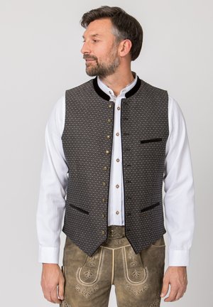 Mann trägt traditionelle bayerische Kleidung mit weißem Hemd, gemusterter dunkler Weste und braunen bestickten Lederhosen, schaut zur Seite.