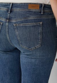 Jeans dżinsowe w ciemnoniebieskim odcieniu o gładkiej teksturze. Cechy to dwa tylne kieszenie oraz brązowa, sztuczna skórzana metka z logo na pasku.
