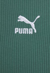 Logo Puma blanc avec un chat sautant et du texte sur un fond en tissu côtelé vert.