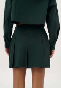 Donkergroene geplooide rok met een gladde textuur, met een strakke tailleband en een cropped top die gedeeltelijk zichtbaar is aan de achterkant.