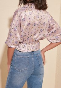 Blouse en satin de couleur rose pastel avec un motif floral violet et jaune, à manches courtes et taille froncée. Portée avec un jean bleu taille haute.