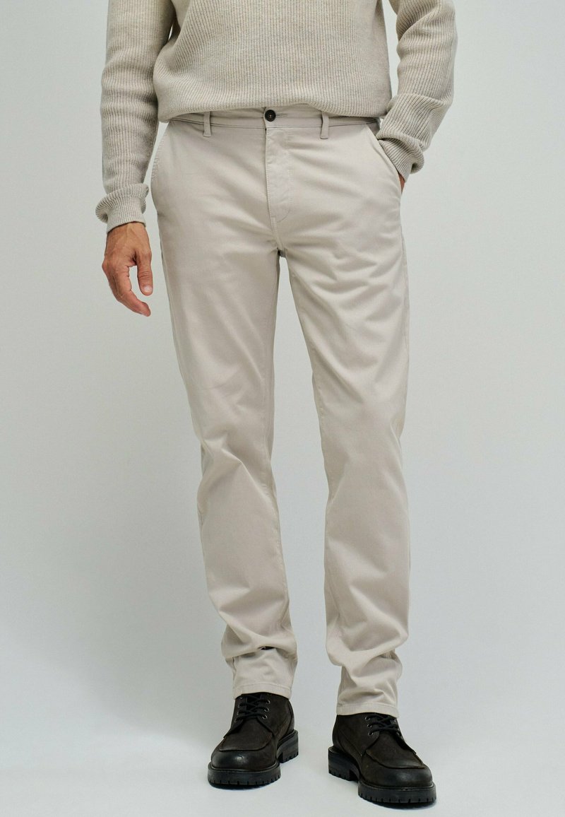 Mężczyzna w beżowych spodniach chino, beżowym prążkowanym swetrze, czarnych sznurowanych butach, stojący z jedną ręką w kieszeni na jasnym tle.