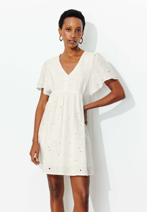 Femme portant une robe courte blanche en œillet avec des manches volantées et un décolleté en V, debout avec une main sur la hanche sur fond blanc.