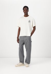 Camiseta blanca de manga corta con un pequeño gráfico oscuro; pantalones grises a rayas de corte holgado; zapatillas de color crema. Diseño simple con un ajuste casual.