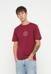 Burgunderfarget bomulls t-skjorte med korte ermer, prydet med et sirkulært logo på brystet. Vist på en modell som har på seg lyseblå jeans.