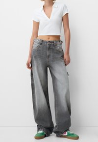 Kobieta w luźnych, wyblakłych szarych dżinsach, skróconej białej koszulce polo i zielonych trampkach na białym tle.